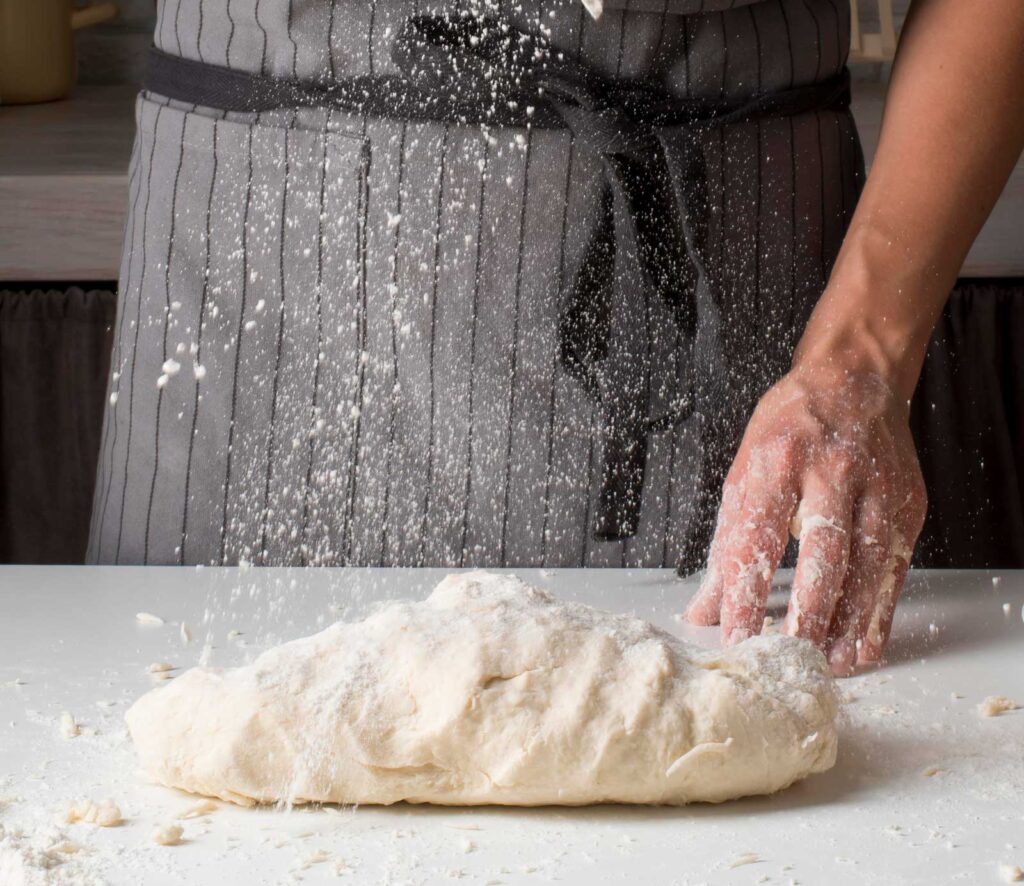 Panificatore al lavoro mentre impasta a mano un impasto rustico su un piano di lavoro infarinato, con strumenti da panificazione e sacchi di farina sullo sfondo. L’immagine cattura l’artigianalità e la passione per la panificazione tradizionale.